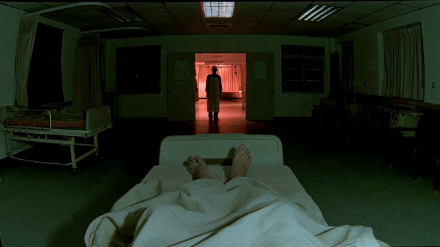 The Surgeon's distant silhouette stands in the red-lit doorway beyond the patient's bed